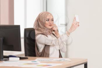 Young Cheerful Muslim Business Woman Working With Computer At Desk In The Modern Office And Taking Selfies With Mobile Phone