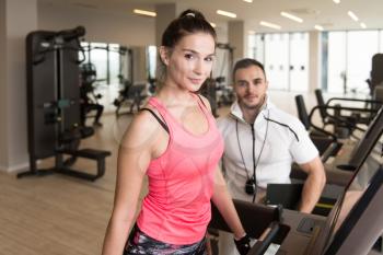Personal Trainer Showing Young Woman How To Train Aerobics Elliptical Walker In The Gym