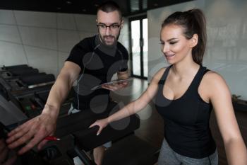 Personal Trainer Showing Young Woman How To Train Aerobics Elliptical Walker In The Gym