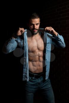 Portrait Of A Young Physically Fit Man Showing His Well Trained Body While Wearing Blue Shirt - Muscular Athletic Bodybuilder Fitness Model Posing After Exercises On Wall of Bricks