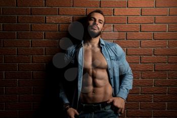 Portrait Of A Young Physically Fit Man Showing His Well Trained Body While Wearing Blue Shirt - Muscular Athletic Bodybuilder Fitness Model Posing After Exercises On Wall of Bricks