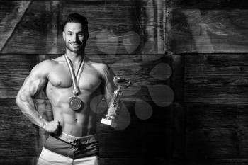 Bodybuilder Competitor Showing His Winning Medal Against a Wooden Wall and Flexing Muscles While Wearing Sports Shorts - Male Fitness Competitor Showing His Winning Medal a Place for Your Text