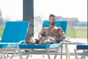 Young Man Resting On Sun Loungers By Swimming Pool And Flexing Muscles