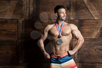 Athlete Competitor Showing His Winning Medal Against a Wooden Wall and Flexing Muscles While Wearing Sports Shorts - Male Fitness Competitor Showing His Winning Medal a Place for Your Text