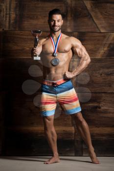 Athlete Competitor Showing His Winning Medal Against a Wooden Wall and Flexing Muscles While Wearing Sports Shorts - Male Fitness Competitor Showing His Winning Medal a Place for Your Text