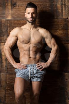 Portrait of a Young Physically Fit Man Showing His Well Trained Body While Wearing Sports Shorts - Muscular Athletic Bodybuilder Fitness Model Posing After Exercises on Wooden Wall - a Place for Your Text