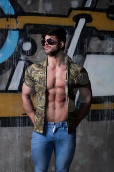 Portrait of a Young Physically Fit Man in Army Shirt Showing His Well Trained Body - Muscular Athletic Bodybuilder Fitness Model Posing After Exercises In Front Of A Graffiti Wall