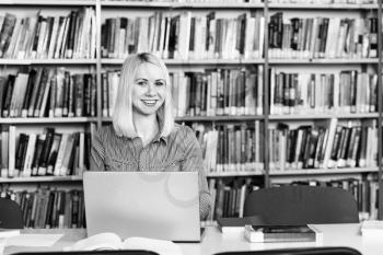 Pretty Woman With Blonde Hair Sitting at a Desk in the Library - Laptop and Organiser on the Table - Looking at the Screen a Concept of Studying - Blurred Books at the Back