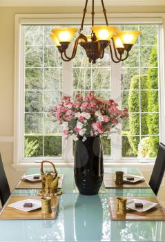 Vertical photo of dining room table with flowers, tea pot, tea cups, white plates with cookies on bamboo placemats in family formal dining room with daylight coming through windows in background