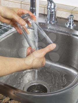 Vertical photo of female hands drying off a drinking glass with water running from kitchen faucet sink in background