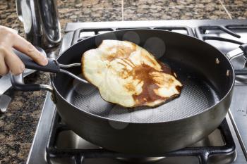 Horizontal photo of main focus on pancake being flipped in hot frying pan on top of gas stove range