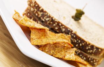 Horizontal photo of golden chips in front of whole wheat sandwich on white plate with natural bamboo wood underneath