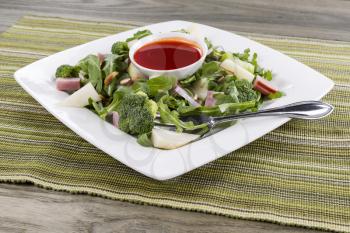 Freshly made green salad with salad dressing in small bowl and fork on white square plate