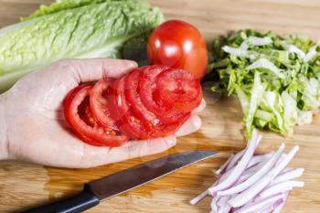 Horizontal photo of female hand with sliced tomato in hand, lettuce, onion on natural bamboo cutting board