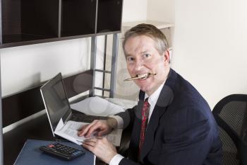 Mature man chewing pen while working on income tax with calculator, computer and papers on desk