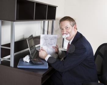 Mature man chewing paper while holding papers in hand with calculator and computer on desk