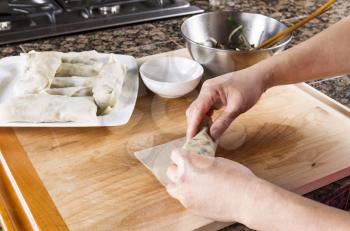 Horizontal photo of Female hands with focus on spring roll being assembled with plate full of finish rolls and kitchen stove in background