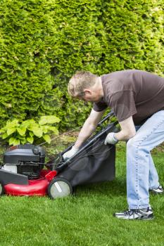 Vertical photo of a mature man, dressed in blue jeans and t-shirt, putting grass bag on old gas lawnmower with tall bushes in background