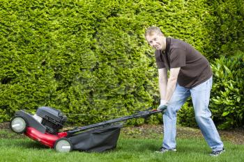 Horizontal photo of a mature man, dressed in blue jeans and t-shirt, while getting ready to use an old gas lawnmower with tall bushes in background