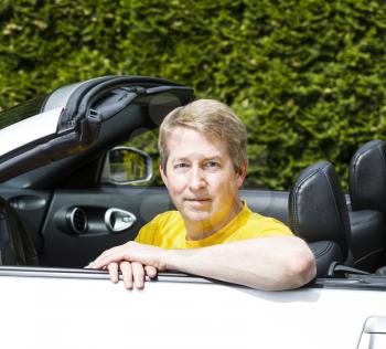 Horizontal photo of a mature man sitting in a convertible car with arm on top of door, while wearing a yellow short sleeve shirt, outdoors on a sunny day