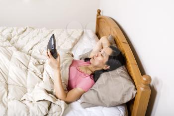 Horizontal photo of mature woman, lying head down in pillow, while looking at her alarm clock in her hand