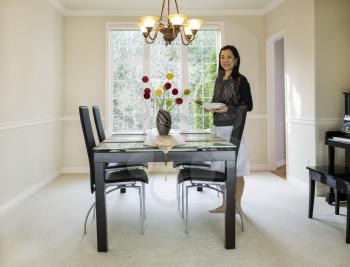 Photo of mature woman placing diner plates and chop sticks  in family formal dining room with daylight coming through large windows in background