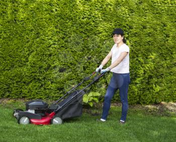 Horizontal photo of a mature woman preparing to use an old gas lawnmower on grass yard with tall bushes and flower garden in background