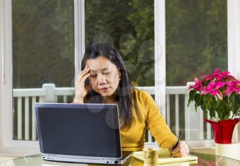 Mature Asian woman working at home, looking stressed, with notebook computer on glass table with large windows in background