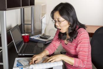 Mature Asian woman surprised while holding  income tax tables booklet with computer, coffee cup and calculator on desk