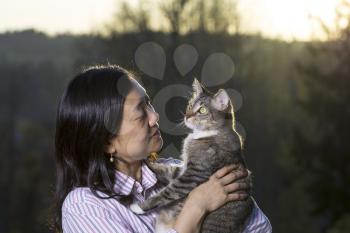 Horizontal photo of mature woman holding cat outdoors with fading sunset and trees in background