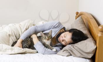 Horizontal photo of mature woman, lying head down in pillow, while hold her family cat in bed