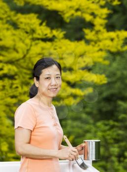 Vertical photo of a mature woman holding paint can and brush while preparing to work on white wood deck railing with a green nature background
