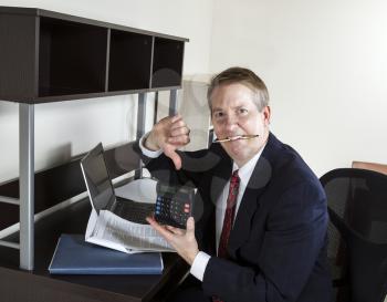 Mature man chewing pen while pointing thumbs down on calculator, with computer and papers on desk