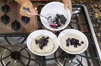 Blueberries being added to Oatmeal Breakfast in white bowls on stove top
