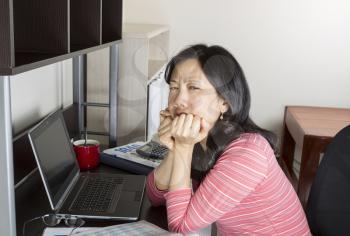 Mature Asian women frustrated doing personal income taxes with computer, coffee cup, calculator and tax booklet on desk