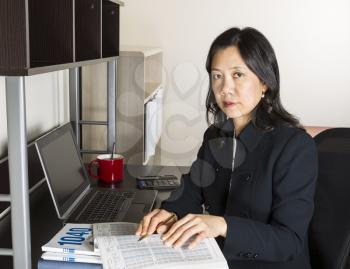 Professional Mature Asian woman working on Personal Income Taxes with tax table booklet, computer, coffee cup with spoon on desk