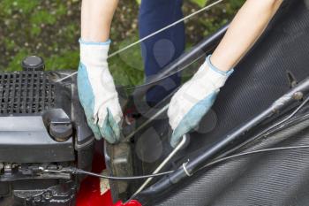 Horizontal photo of a female hands, wearing gloves, while installing catching bag in old gas lawnmower on grass yard with partial jeans and green grass in background