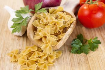 Closeup Horizontal photo of raw pasta in wooden spoon surround by onion, parsley, garlic and tomatoes on natural bamboo board