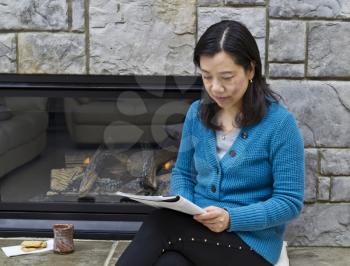 Asian woman sitting next to fireplace reading magazine with coffee cup next to her