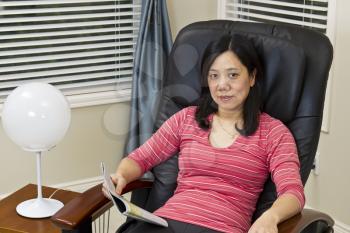 Asian lady reading magazine while relaxing in massage chair at home