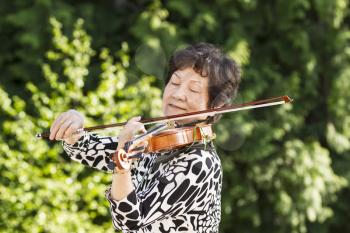 Horizontal photo of Senior Asian woman concentrating while playing the violin outdoors with bright green trees in background