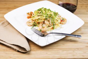 Closeup horizontal photo of Pasta dish with large shrimp, basil, parsley, stainless steel fork, red wine and cloth napkin next to white square plate with natural bamboo wood in background
