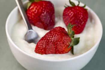 Horizontal photo of a fresh whole strawberries in bowl of vanilla yogurt with spoon in bowl with light blue table cloth in background