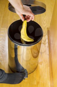 Vertical photo of female putting banana skin into trash can with yellow oak floors in background