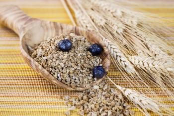 Horizontal photo of wooden spoon filled with Whole Grain Cereal with blueberries on top, wheat stalks on side with yellow table cloth mat in background