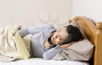Horizontal photo of mature woman, lying head down in pillow, while reading a book in bed