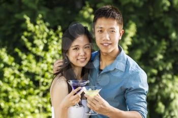 Horizontal photo of a young adult couple, front view,  holding drinks while outdoors with green trees in background