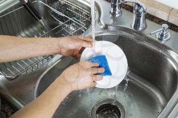 Horizontal photo of female hands washing a small white dinner plate with kitchen sink and running faucet in background