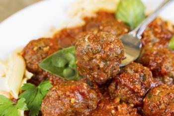 Horizontal photo of bread coated cooked meatball in fork with, pasta, basil, parsley, red sauce and finished meatballs on white plate