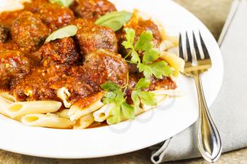 Horizontal photo of bread coated cooked meatballs in pasta with fork, napkin, basil, parsley,and red sauce in round white plate